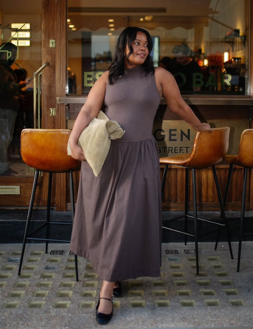 Pessoa com vestido longo marrom, tendência de 2025, em frente a bar com bancos de couro.