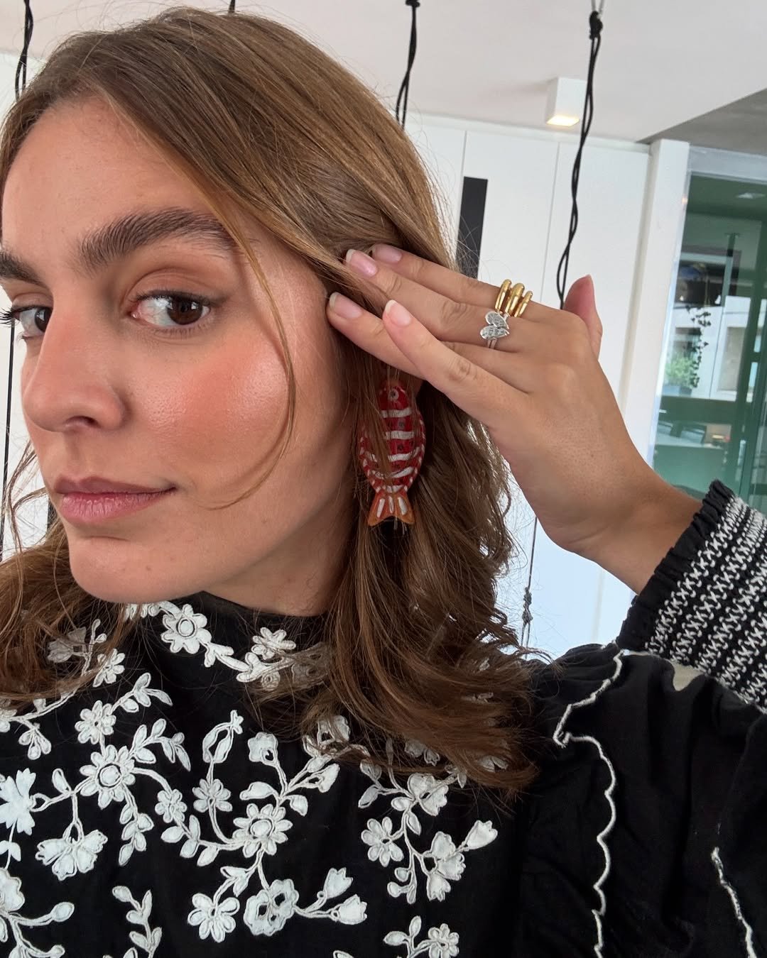 Mulher com sobrancelha grossa exibe brinco de peixe vermelho, com blusa floral preta e branca.