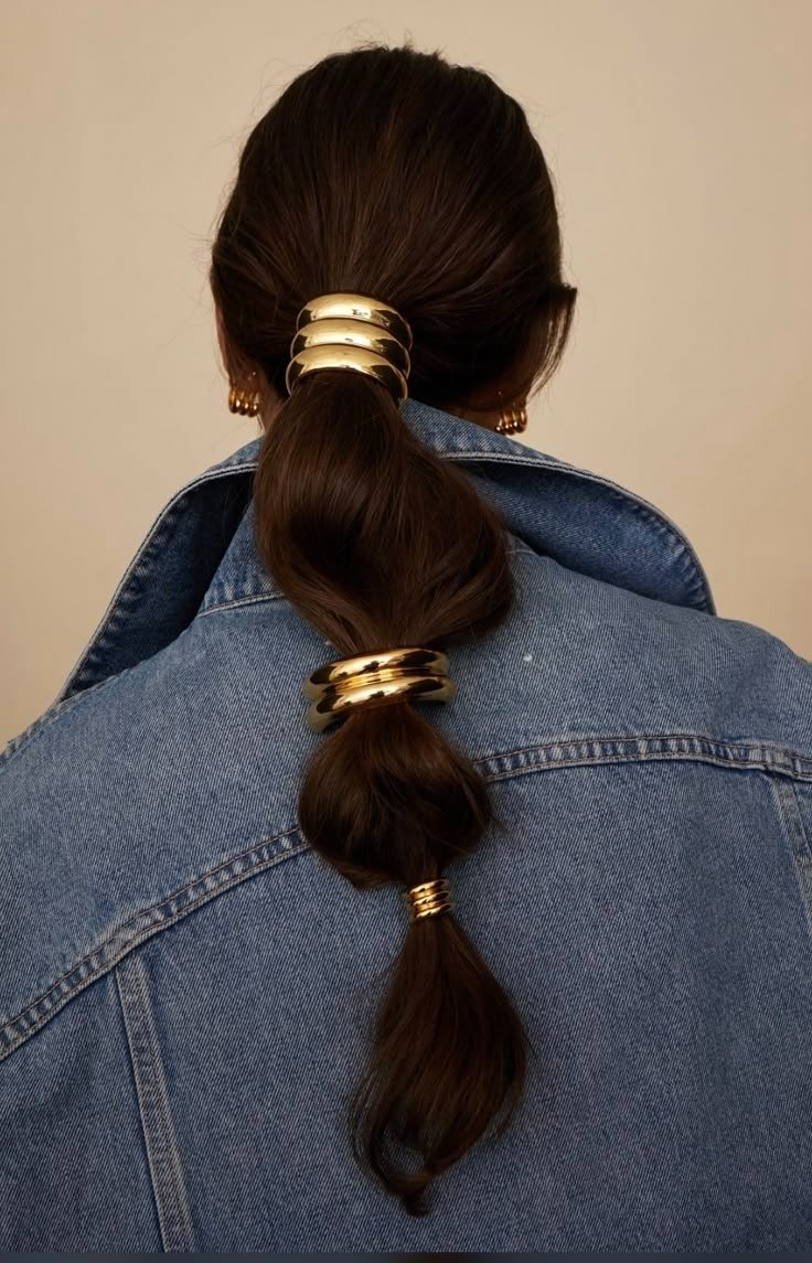 Cabelo preso em rabo de cavalo baixo com acessórios de cabelo chunky dourados, vestido com jaqueta jeans.