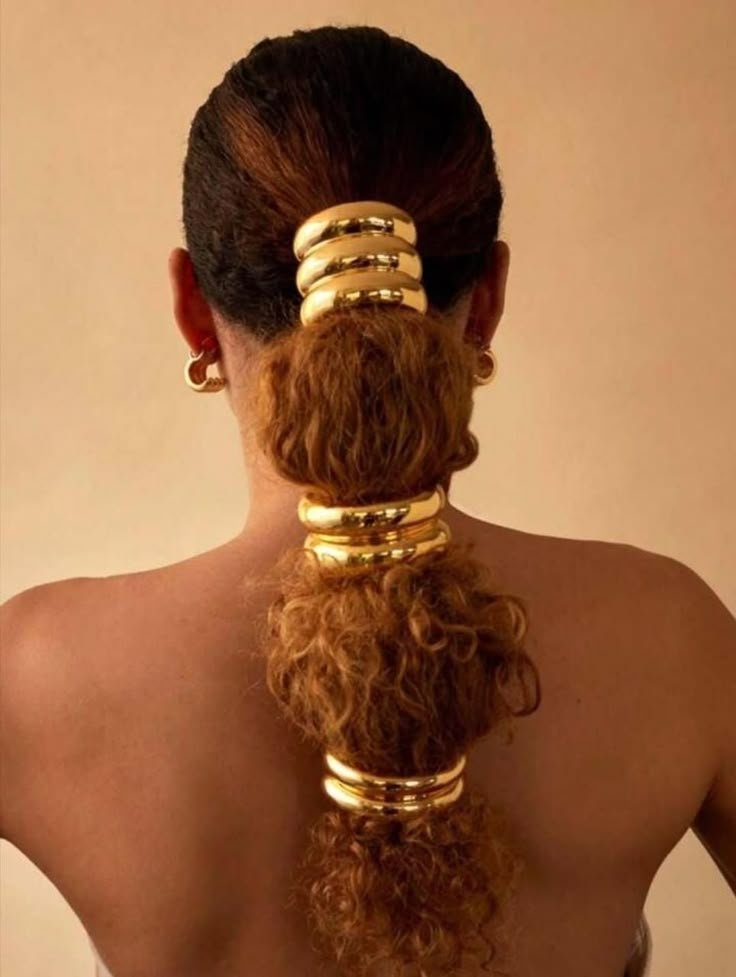 Cabelo cacheado com acessórios dourados grandes, adornando um rabo de cavalo volumoso.