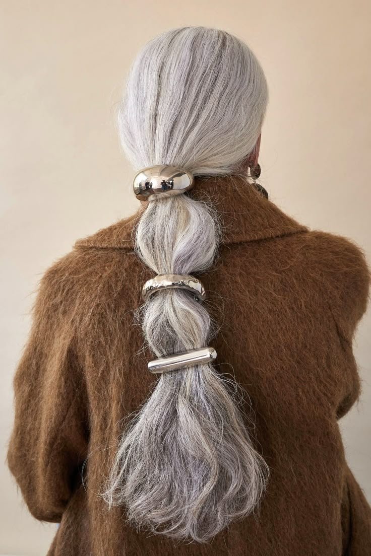 Cabelo cinza preso com acessórios de cabelo chunky prateados, criando um visual moderno sobre um casaco marrom.