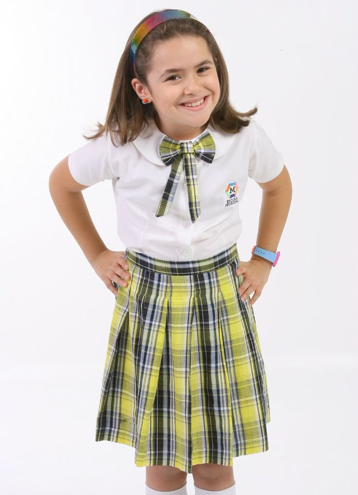 Menina com uniforme escolar xadrez amarelo, sorrindo. Faixa colorida no cabelo. Elemento comum em novelas brasileiras.


