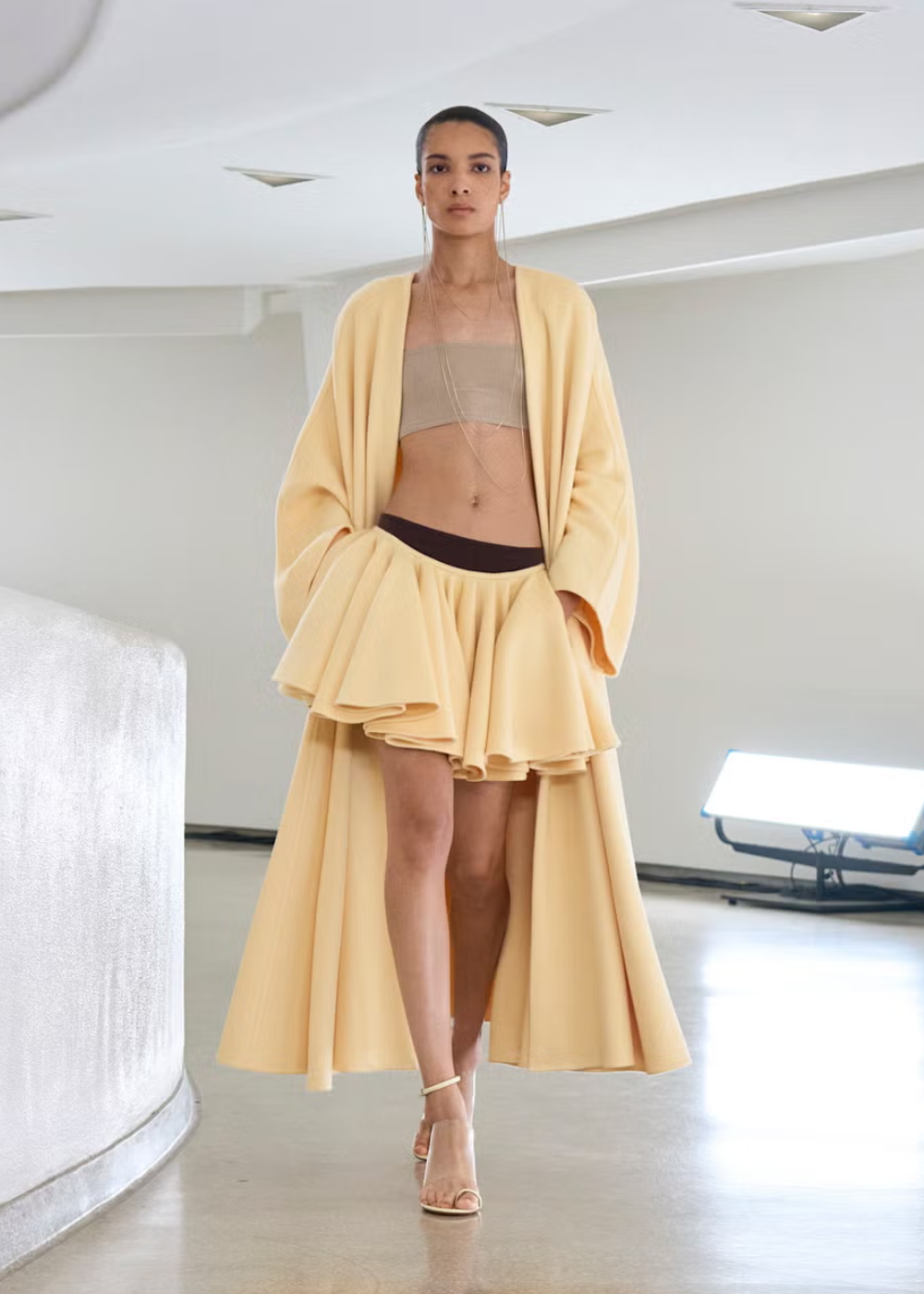 Modelo caminha vestindo conjunto amarelo suave com saia volumosa e casaco longo, em ambiente minimalista.