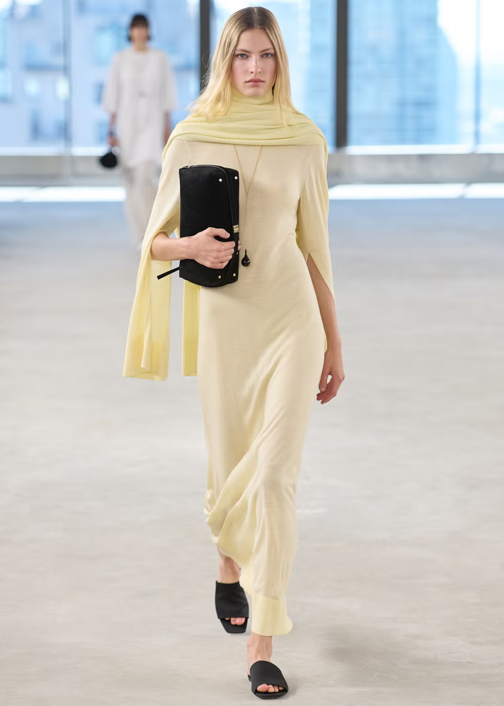 Modelo desfilando com vestido longo em tom amarelo manteiga, carregando bolsa preta.