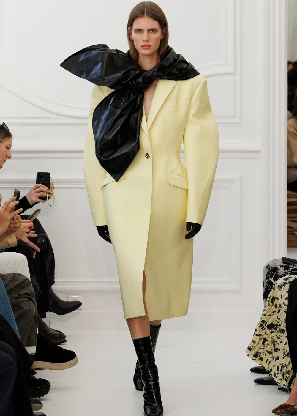 Modelo vestindo casaco amarelo manteiga com laço preto grande, desfilando no inverno.