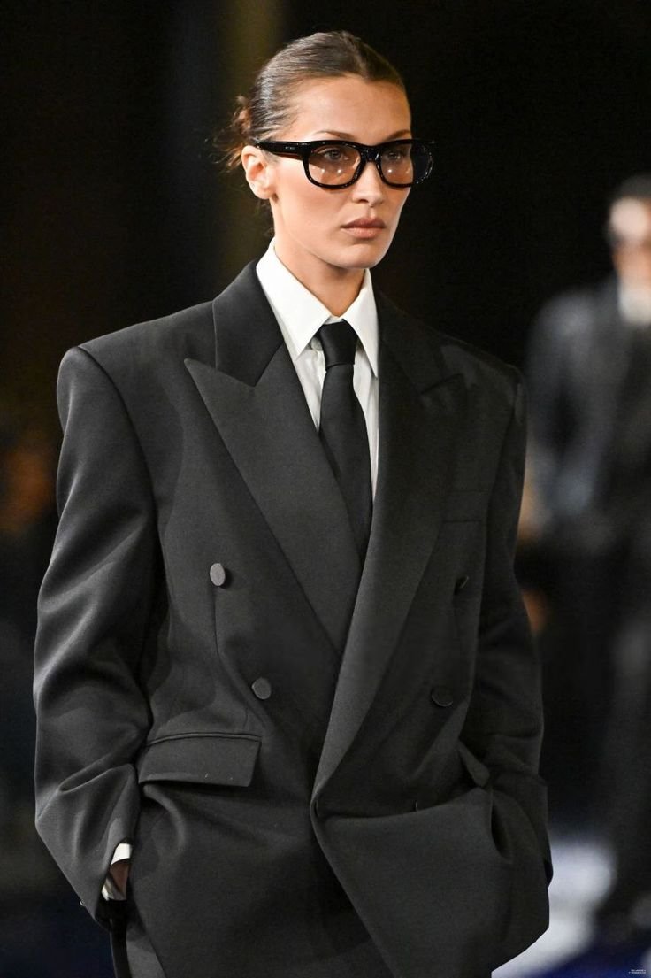 Desfile de moda com modelo usando terninho preto elegante e óculos grandes.
