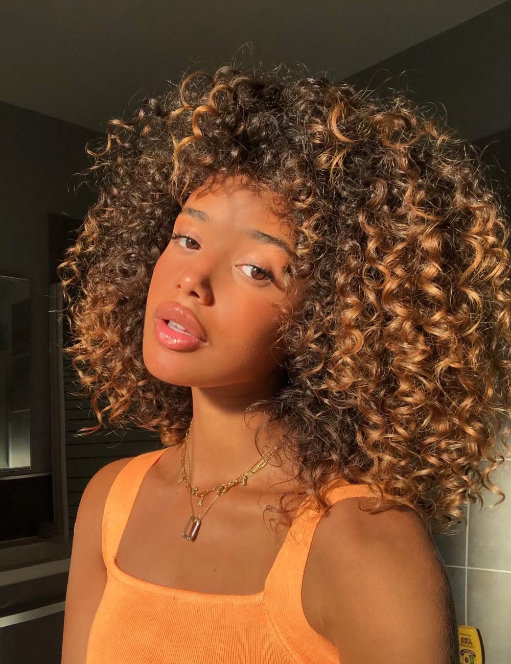 Jovem com cabelos cacheados volumosos e luz solar no rosto, usando blusa laranja.