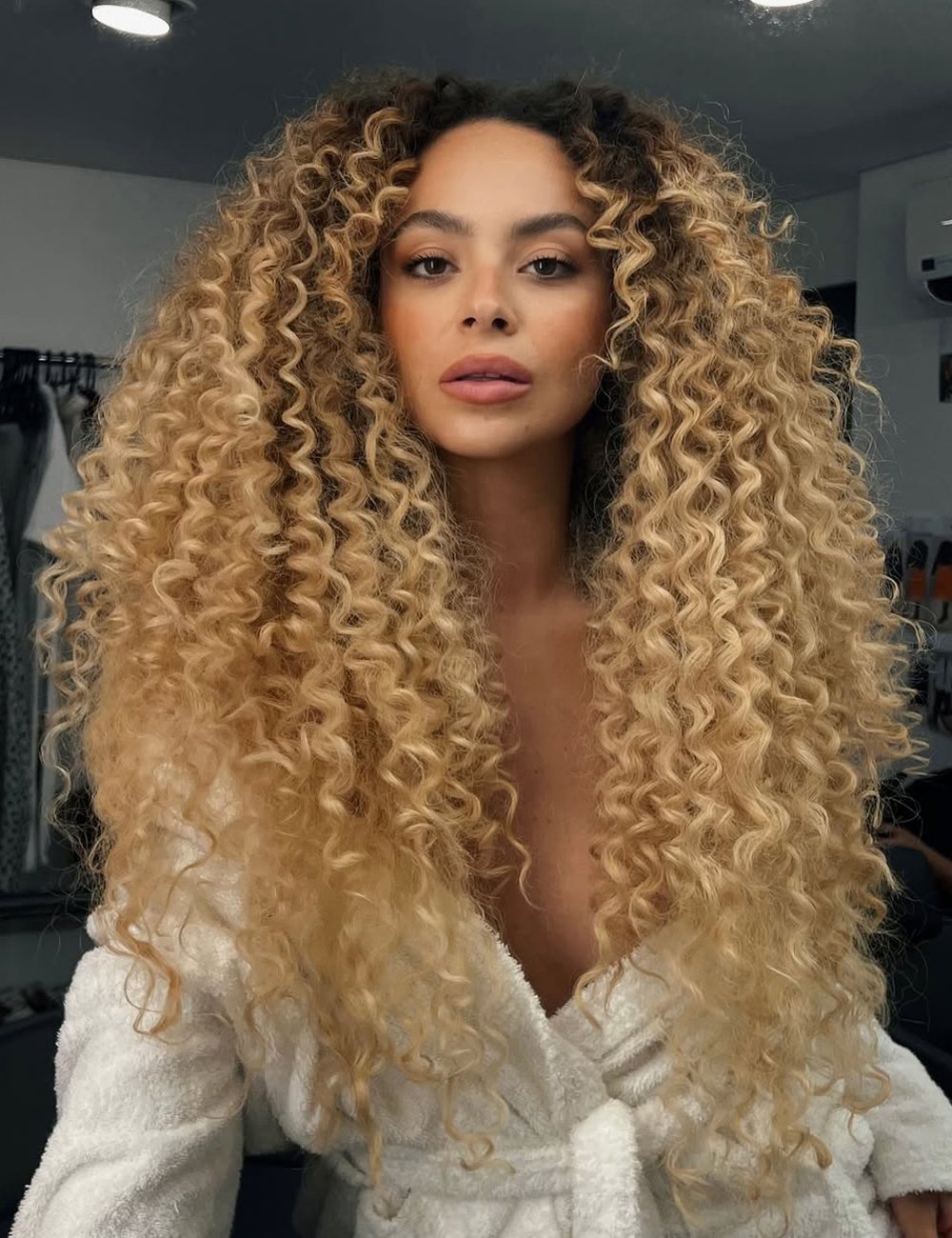 Mulher com cabelos cacheados volumosos e loiros, vestindo roupão branco.