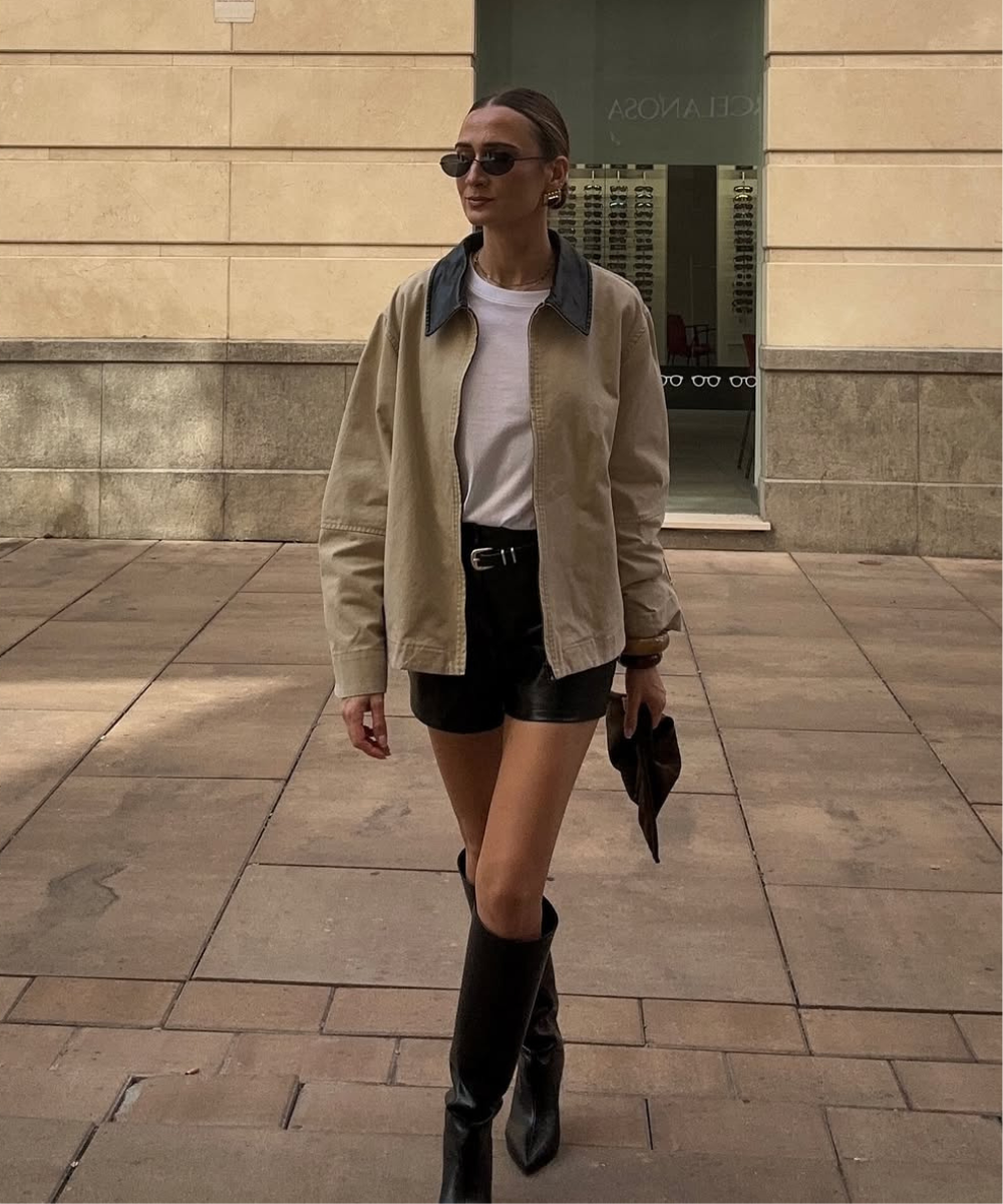 Pessoa andando em calçada, usando casaco bege, camiseta branca, shorts preto e botas, com óculos escuros.