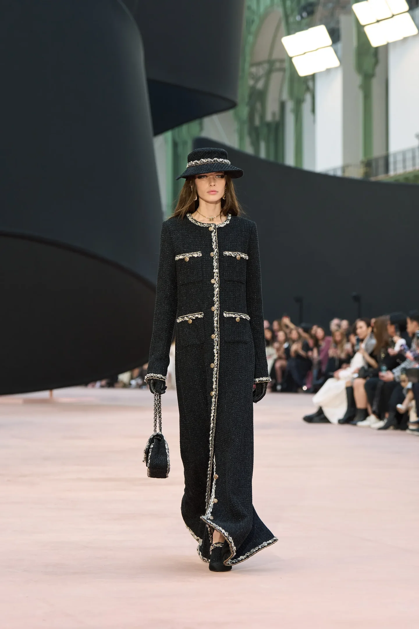 Modelo desfilando em passarela com casaco preto elegante e chapéu combinando. Moda apresenta visual conservador sofisticado.