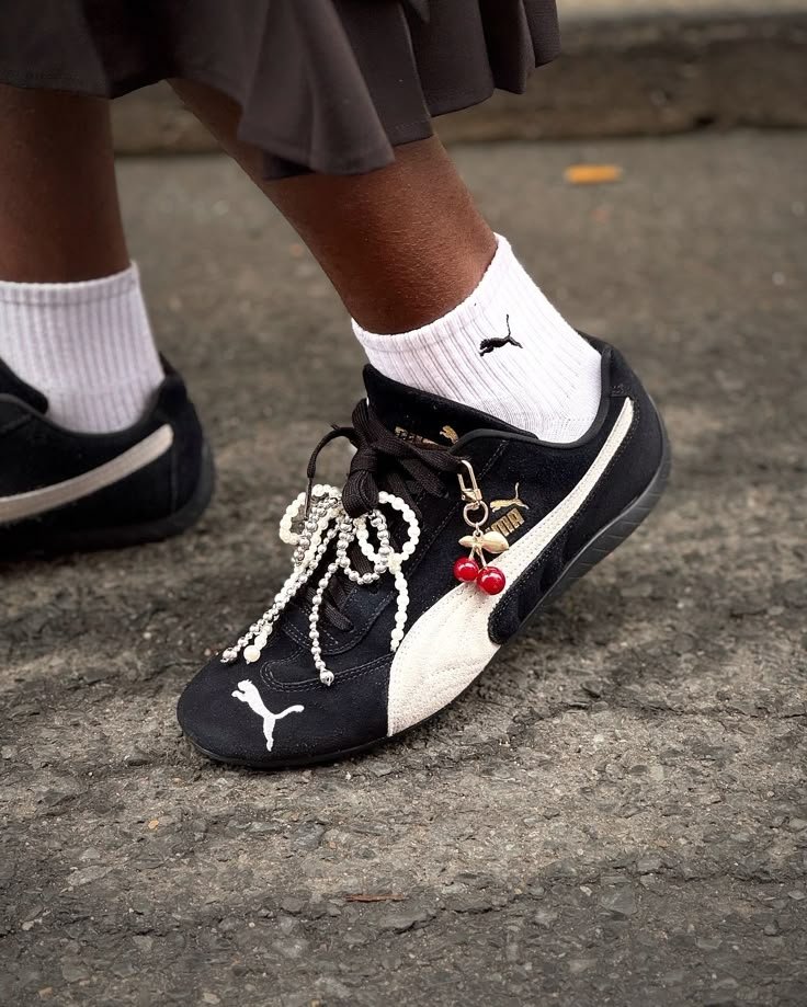 Tênis preto com charms em forma de cereja e pérolas no cadarço, meias brancas e saia escura acima do tornozelo.