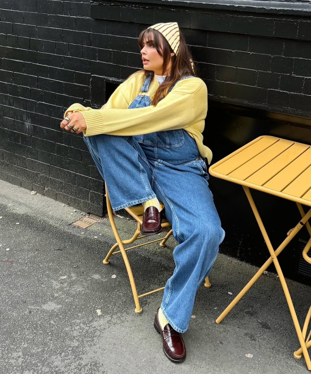 Pessoa sentada em cadeira ao ar livre, usando suéter amarelo manteiga e macacão jeans, com mesa amarela ao lado.