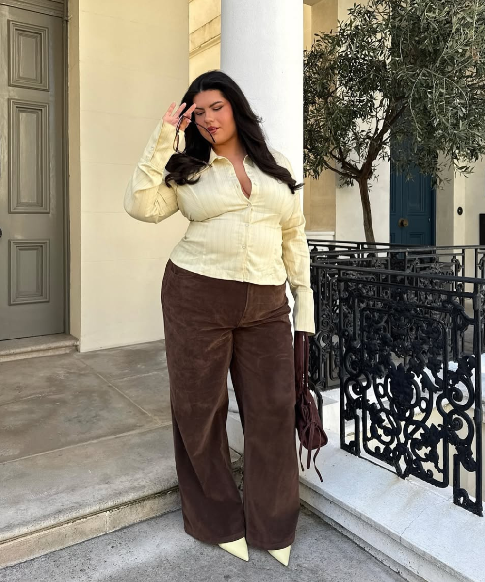 Mulher com camisa amarelo manteiga e calças marrons posa em varanda de inverno elegante.