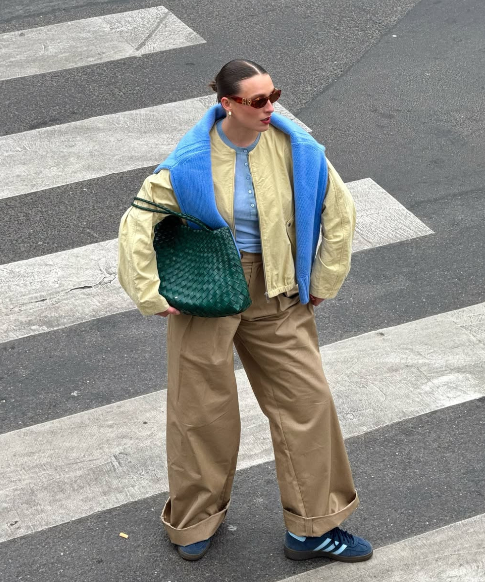 Pessoa usando casaco amarelo manteiga e azul sobre blusa azul, calça bege e óculos marrons, segura bolsa verde.