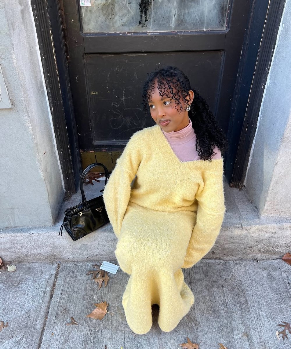 Pessoa em look amarelo manteiga sentada na calçada, clima de inverno. Bolsa preta ao lado.