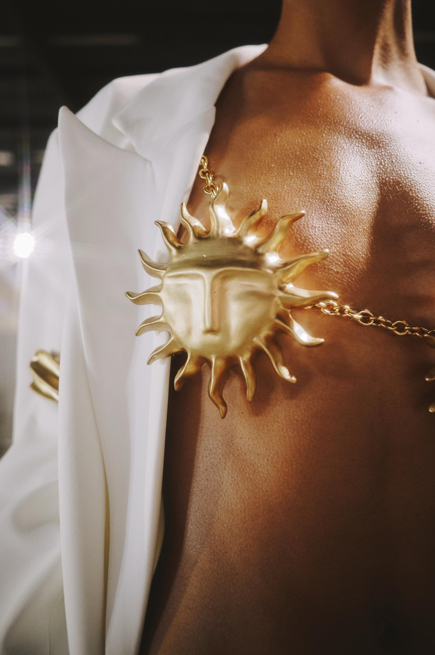 Peça de metal dourado com formato de sol sobre a pele, sob uma jaqueta branca, refletindo tendências de verão.