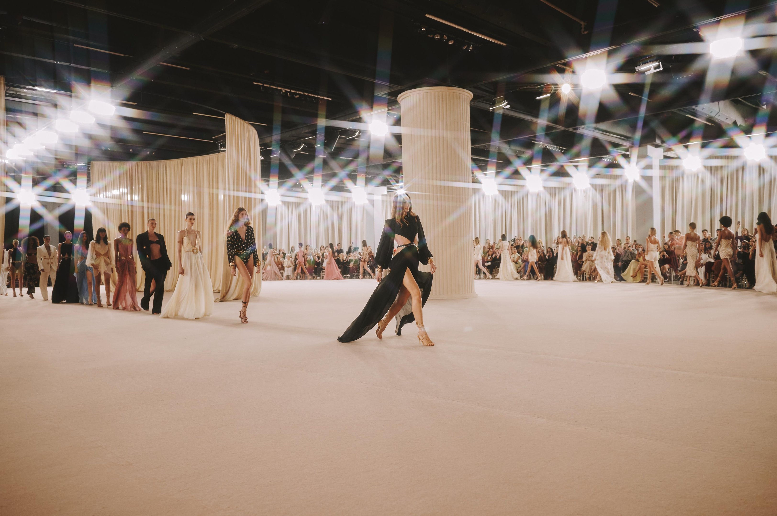 Desfile de moda com modelos exibindo as tendências de verão em trajes elegantes, cercados por colunas e público atento.