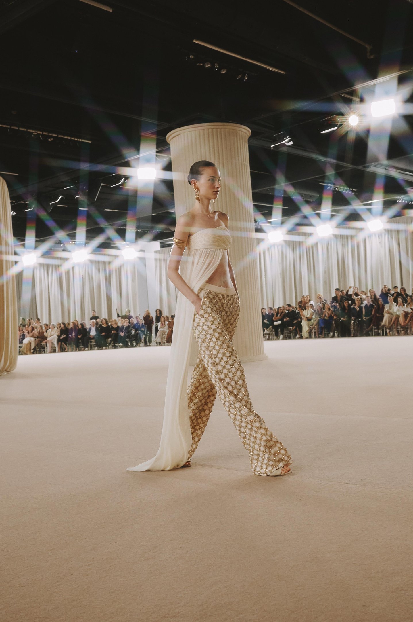 Modelo desfila com calça texturizada e top elegante, refletindo tendências de verão em passarela iluminada.