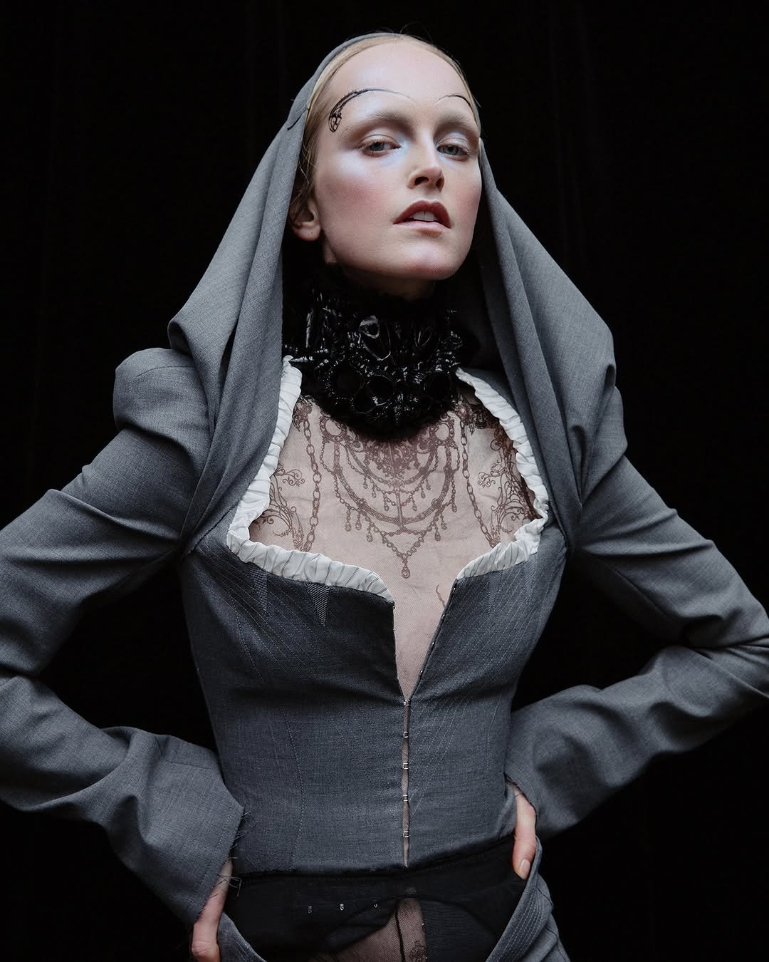Pessoa usando traje cinza com capuz, gola rendada e maquiagem dramática, remetendo a um estilo medieval elegante.