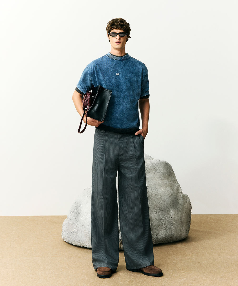Homem com óculos escuros veste camiseta azul e calça larga cinza, segurando bolsa preta. Novidades de moda.