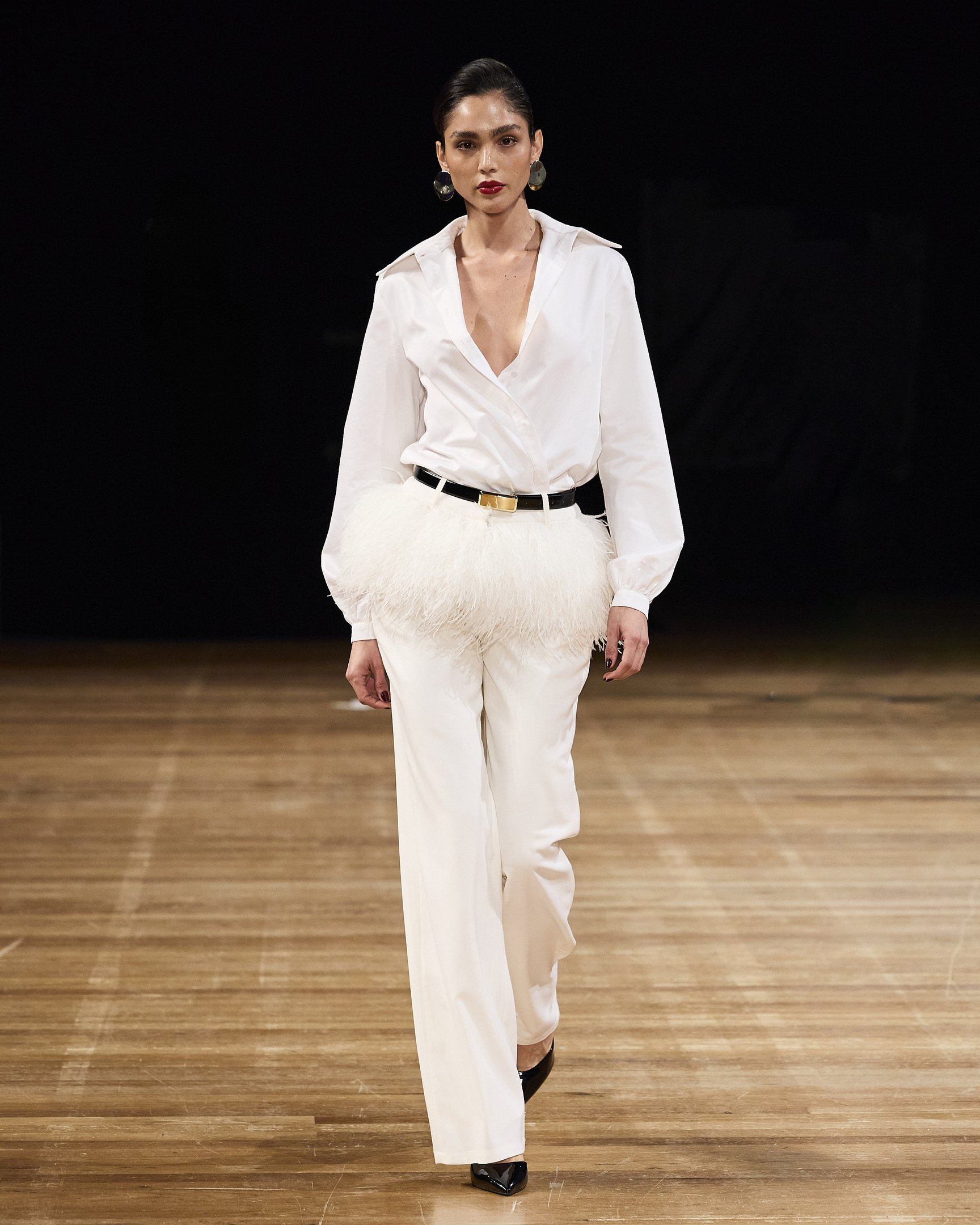 Modelo em passarela usando camisa branca com cinto preto e detalhe de plumas. Fundo escuro. LINI