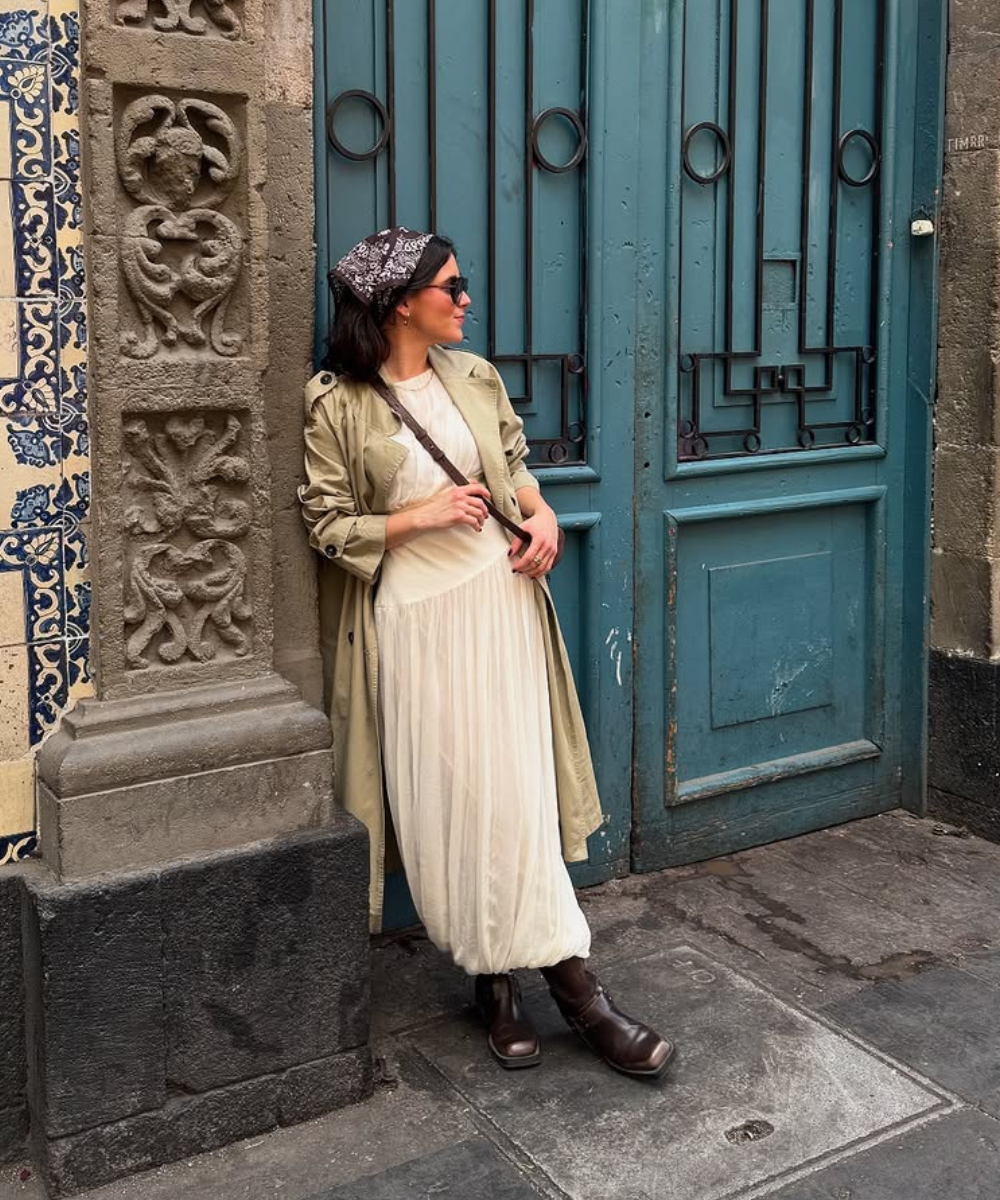 Pessoa com vestido longo e casaco bege, usando botas de cano alto, encostada em uma porta azul.
