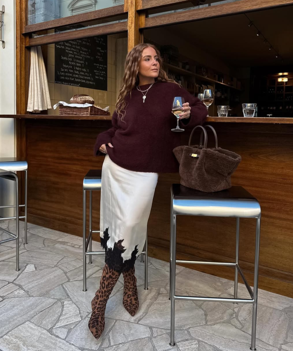 Mulher com suéter vinho e saia clara, usando bota de cano alto estampada, segura uma taça de vinho. Ideia de como usar bota de cano alto com vestidos.