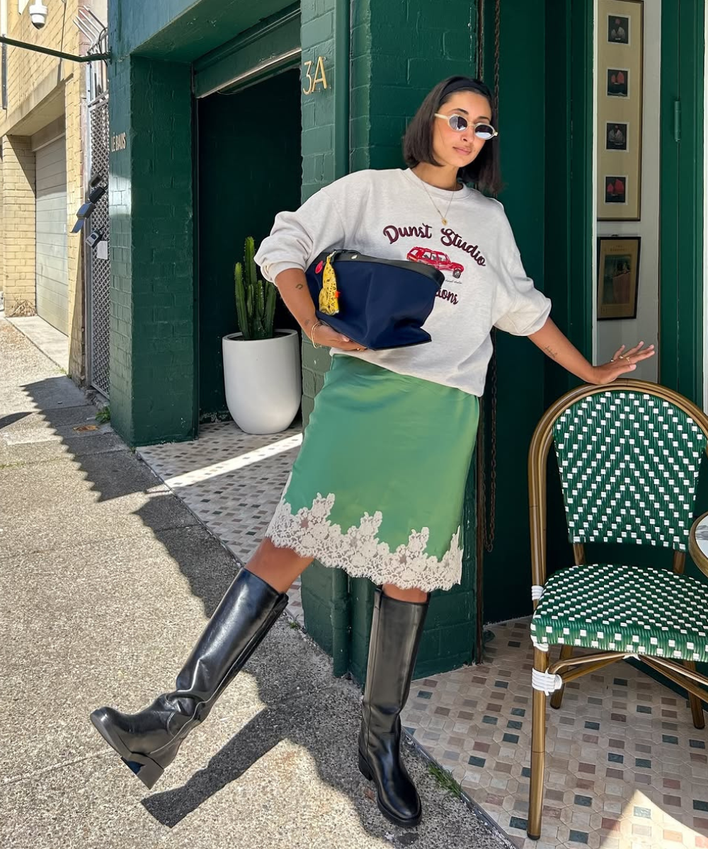 Mulher com moletom e saia verde, usa botas de cano alto ao lado de cadeira verde em ambiente urbano.