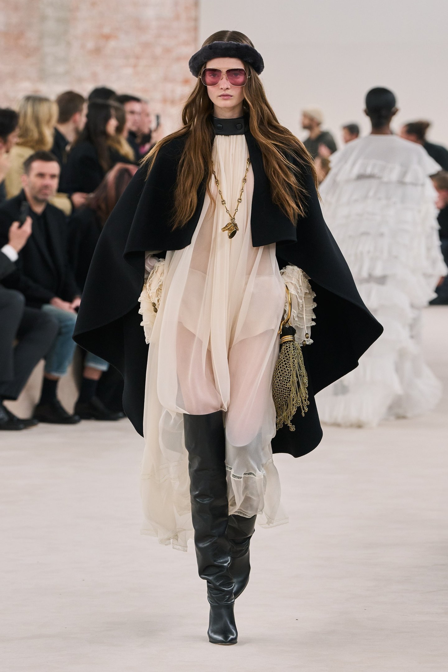 Desfile de moda com looks com bota over the knee. A modelo usa capa preta e vestido claro.