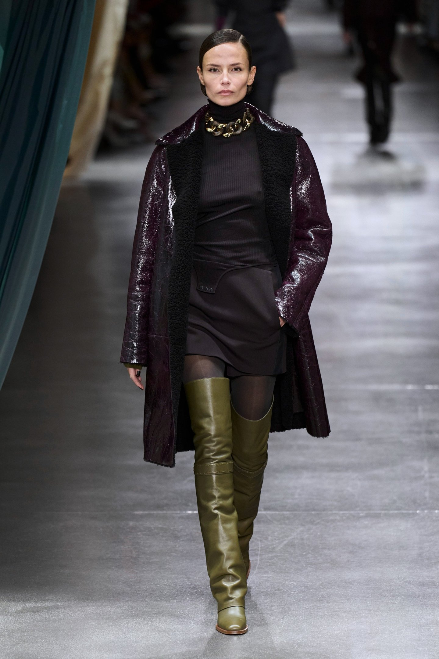 Modelo veste look com bota over the knee verde, casaco vinho brilhante e vestido preto, desfilando em passarela.