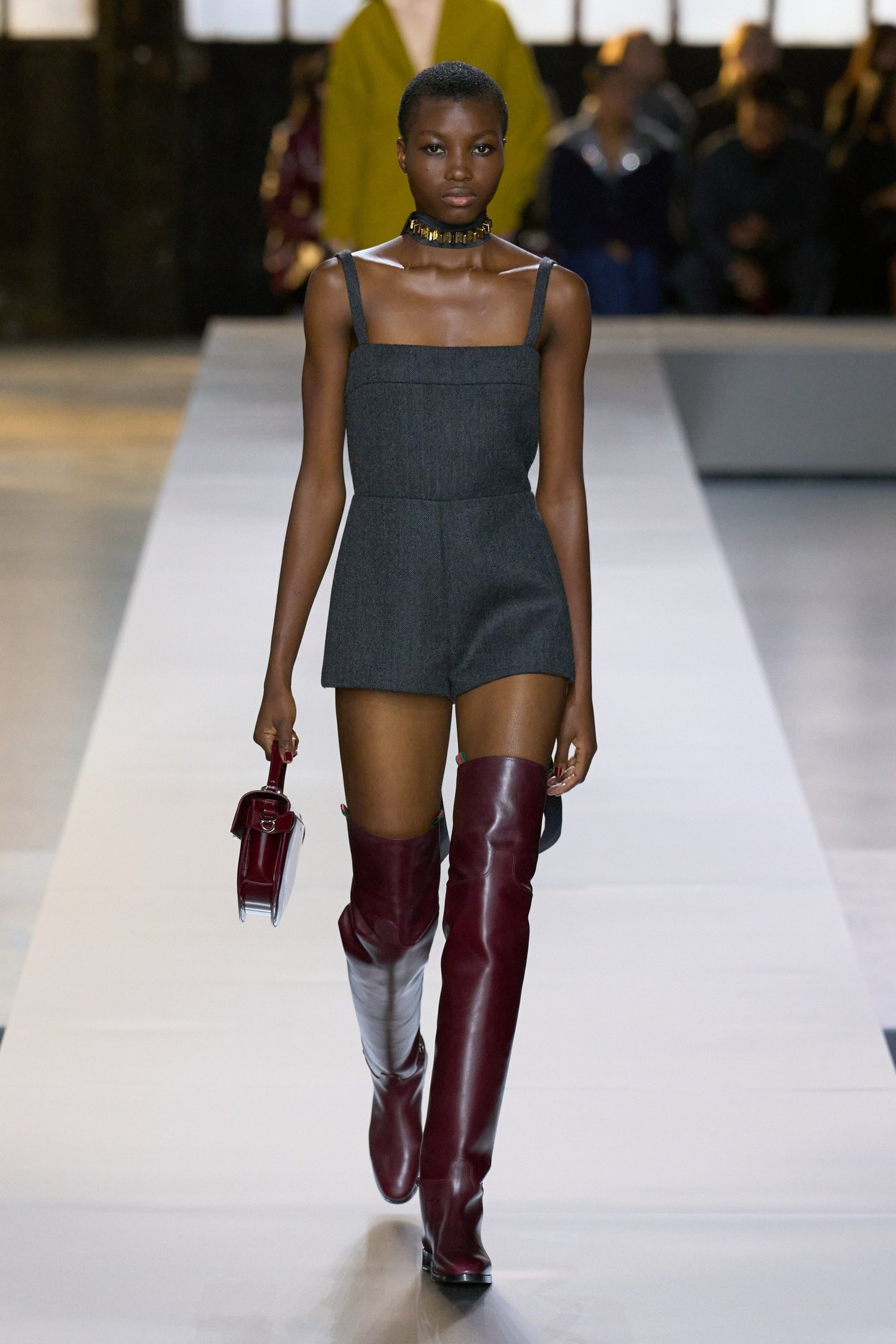 Modelo em passarela usando vestido cinza curto e botas over the knee vinho. O look é complementado por uma bolsa combinando.
