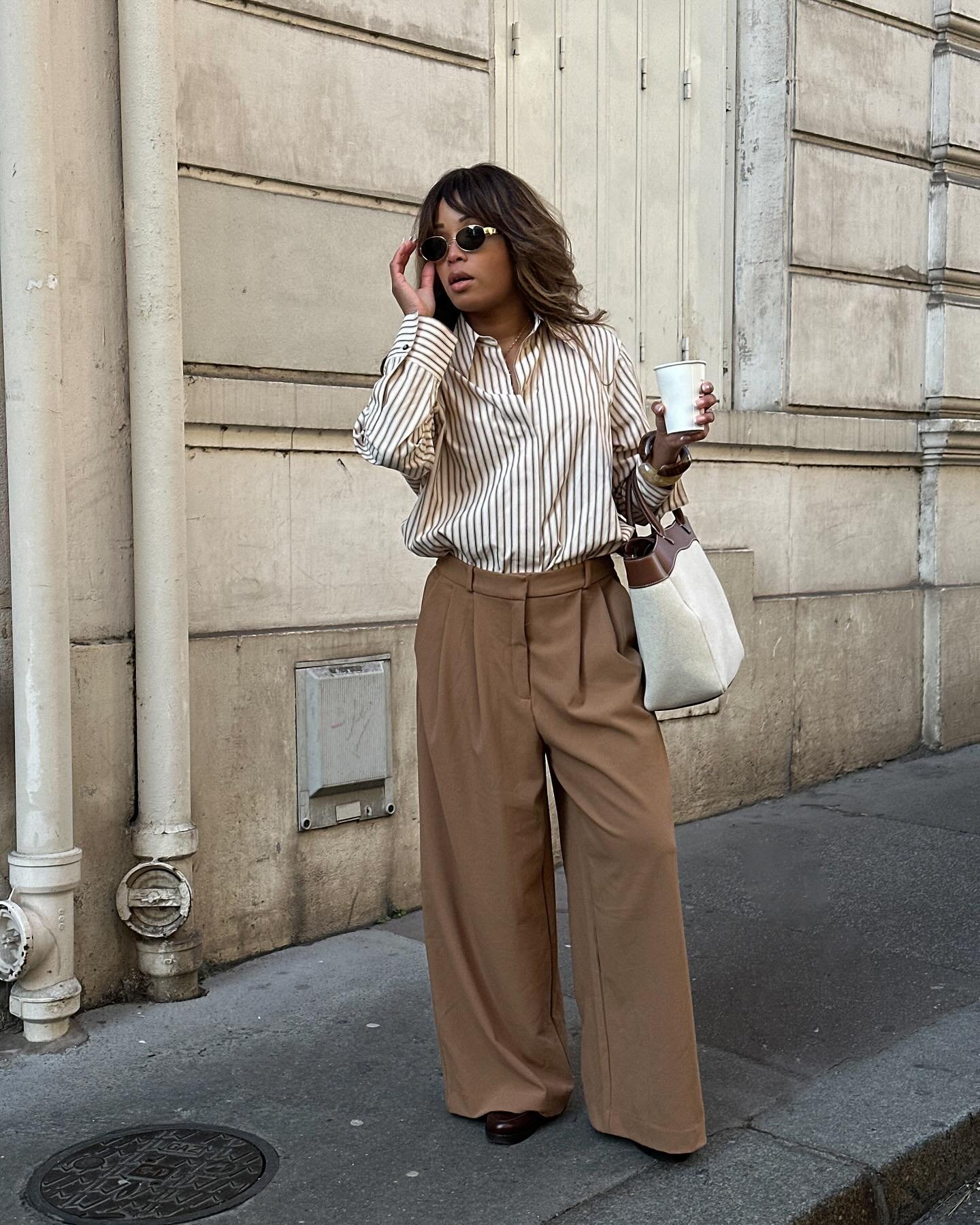 Pessoa usando óculos de sol, camisa listrada e calça wide leg marrom, segurando café e bolsa. Looks com wide leg