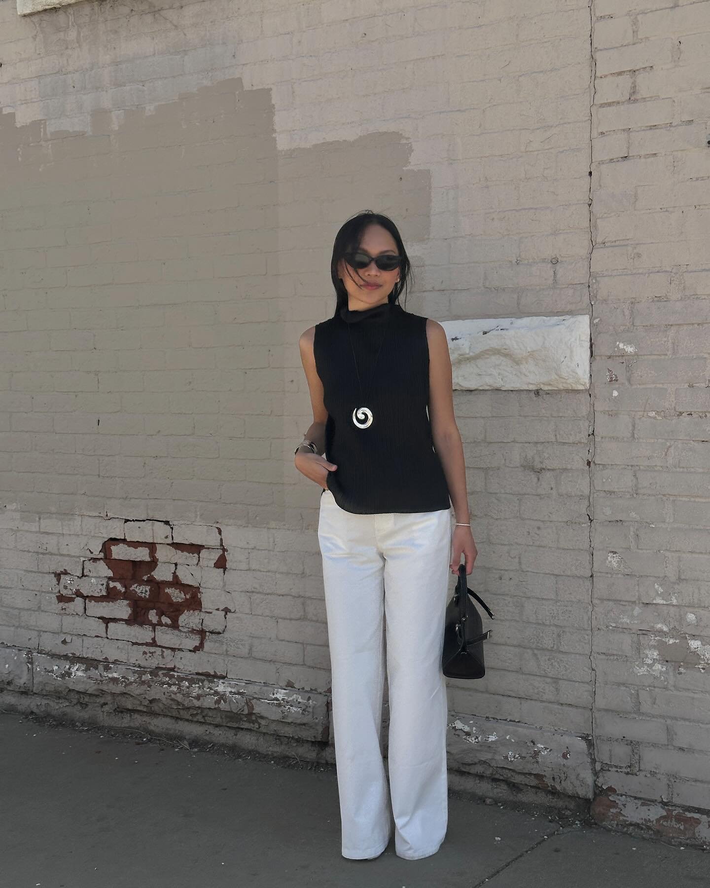 Usuária em look com calça wide leg branca e blusa preta, diante de parede de tijolos desgastados.