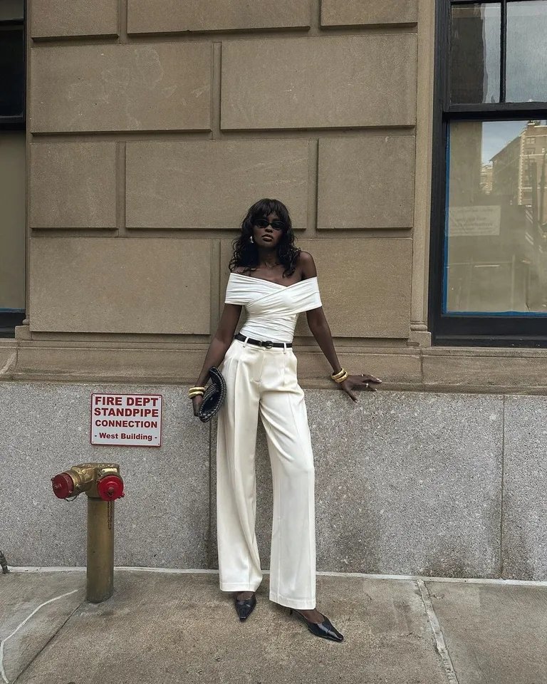 Pessoa posa com top branco e calça de perna larga, ao lado de um hidrante e placa.