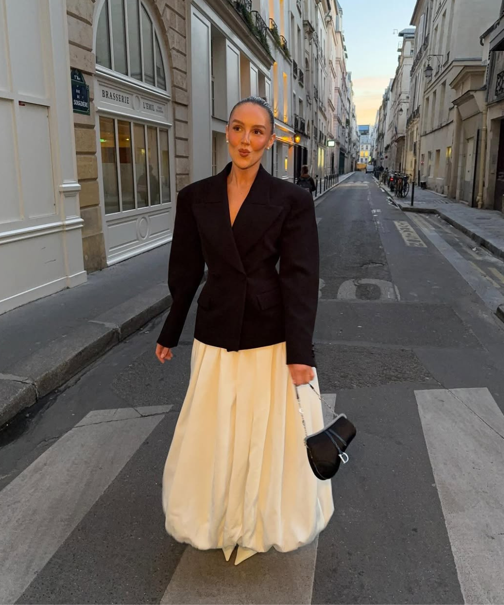 Pessoa com casaco preto e vestido balonê branco em rua de cidade ao entardecer, carregando bolsa preta.