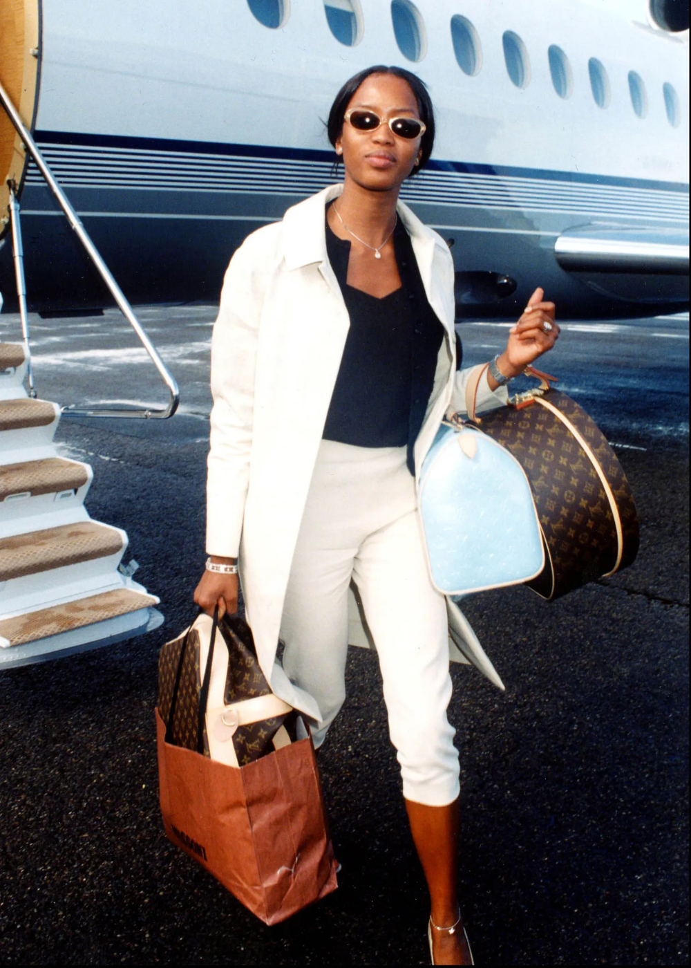 Celebridade com look de aeroporto elegante, carregando bolsas de grife e vestindo casaco claro, óculos de sol.