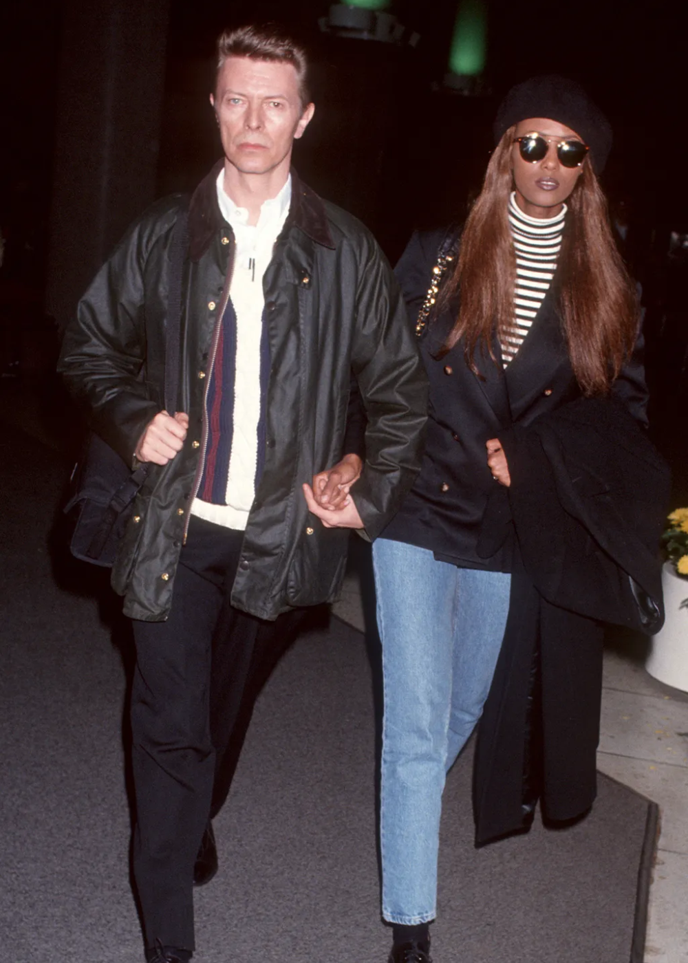 Casal elegante, com ele usando casaco escuro e ela de óculos de sol e boina, exemplificando looks de aeroporto das celebridades.