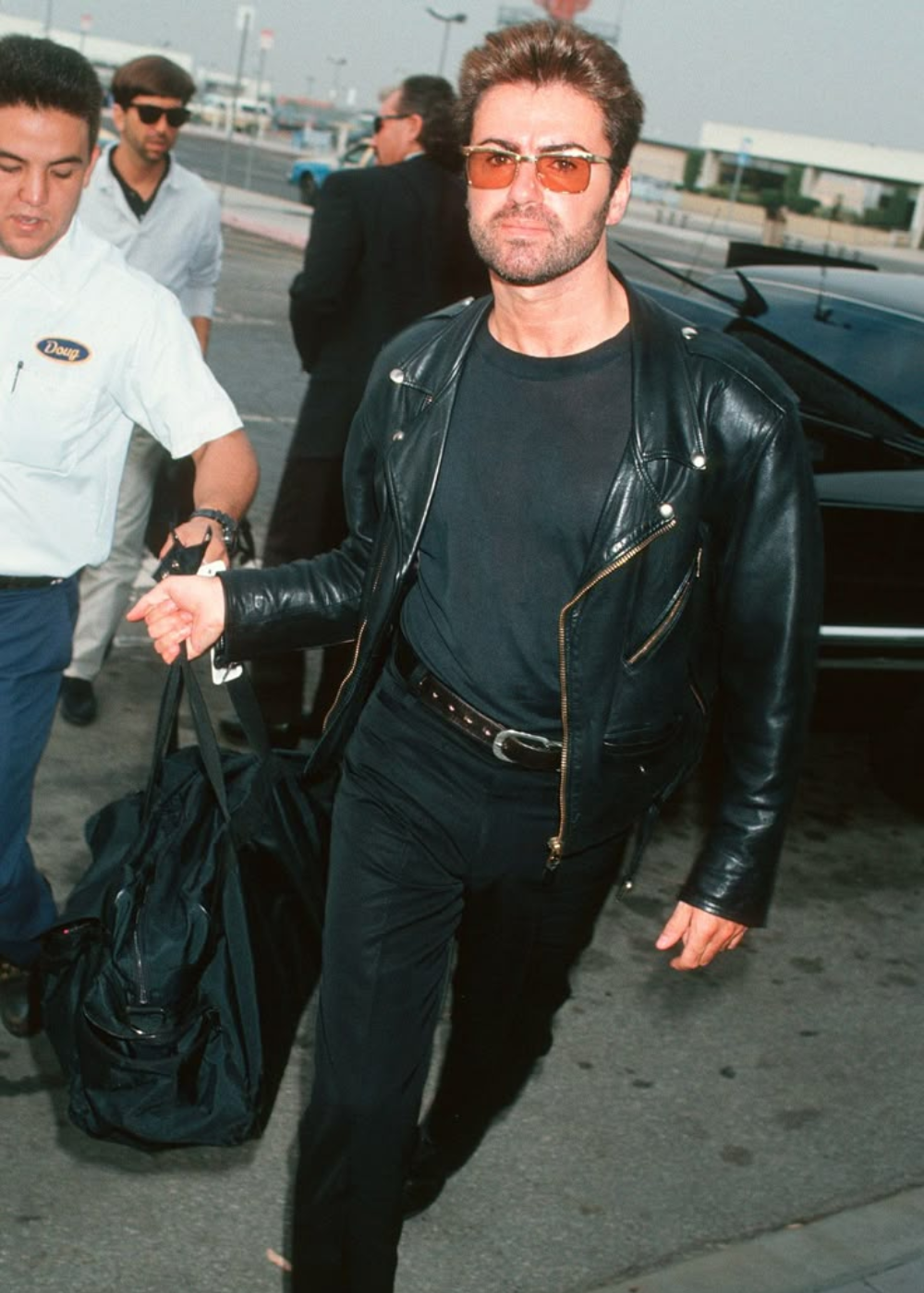 Homem com jaqueta de couro preta e óculos escuros em aeroporto, carregando mala preta. Look casual elegante.