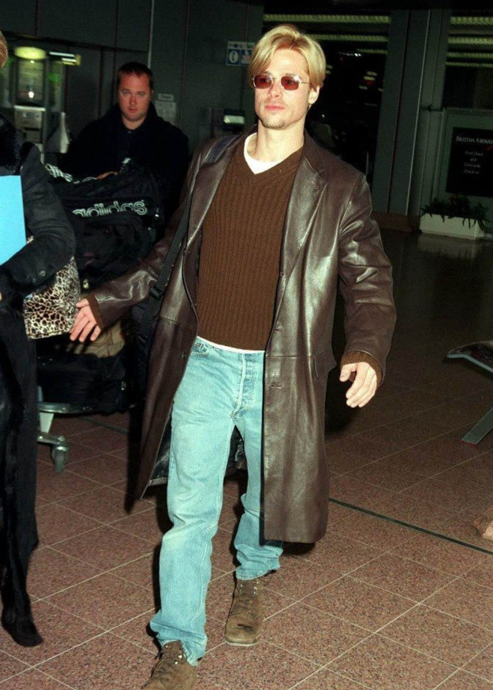 Homem loiro com casaco de couro longo, óculos escuros, suéter marrom e jeans, representando looks de aeroporto das celebridades.
