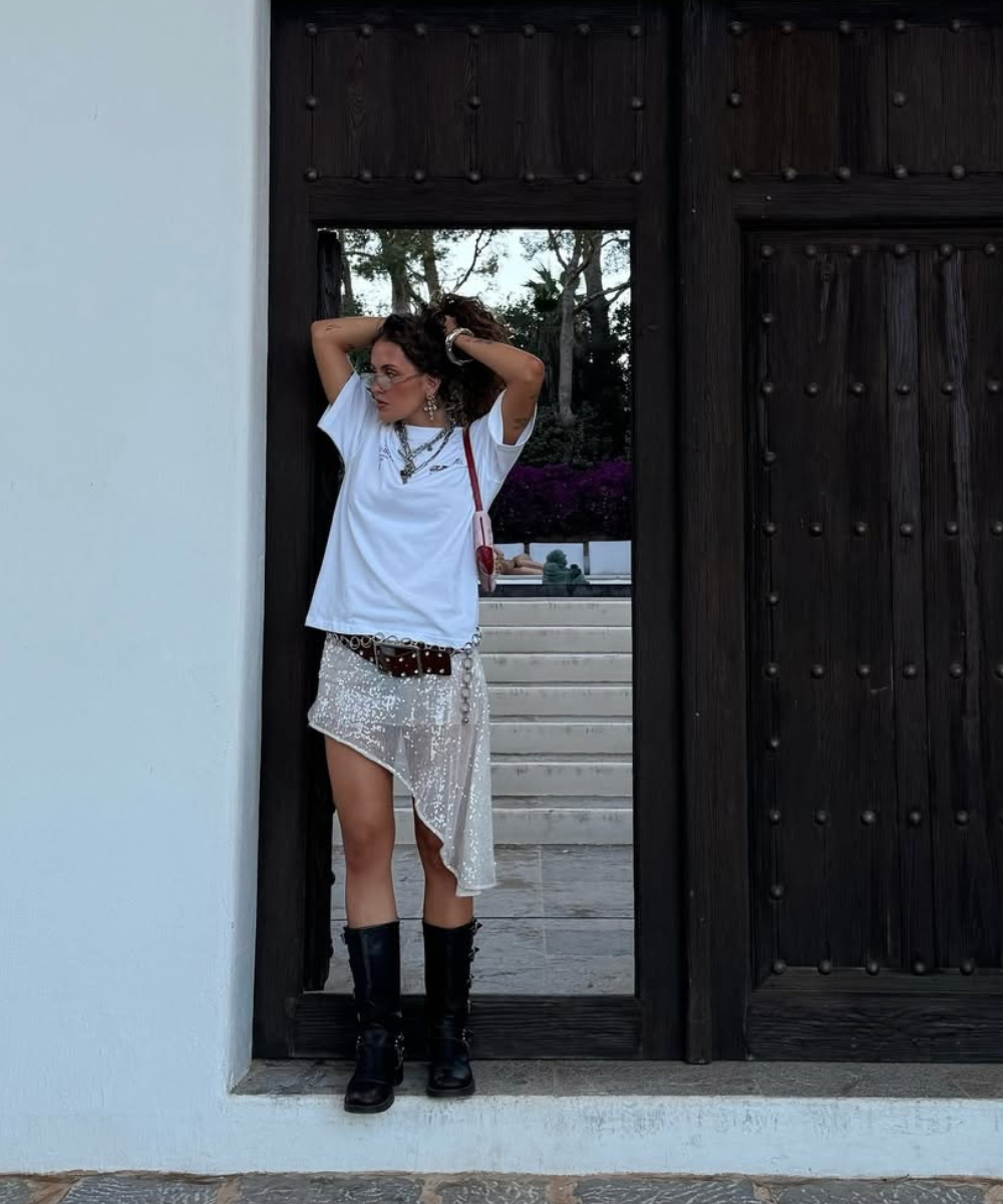 Mulher com saia brilhante, camiseta branca e botas pretas em frente a uma porta de madeira.