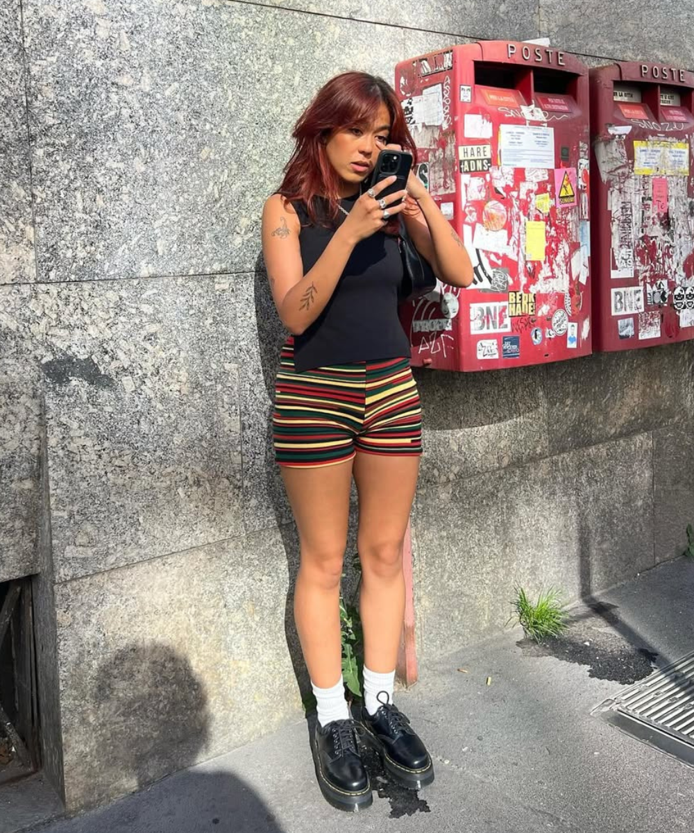 Mulher com shorts listrados usa celular em frente a caixas de correio vermelhas decoradas com adesivos. Inspiração de looks para o show da Lady Gaga.