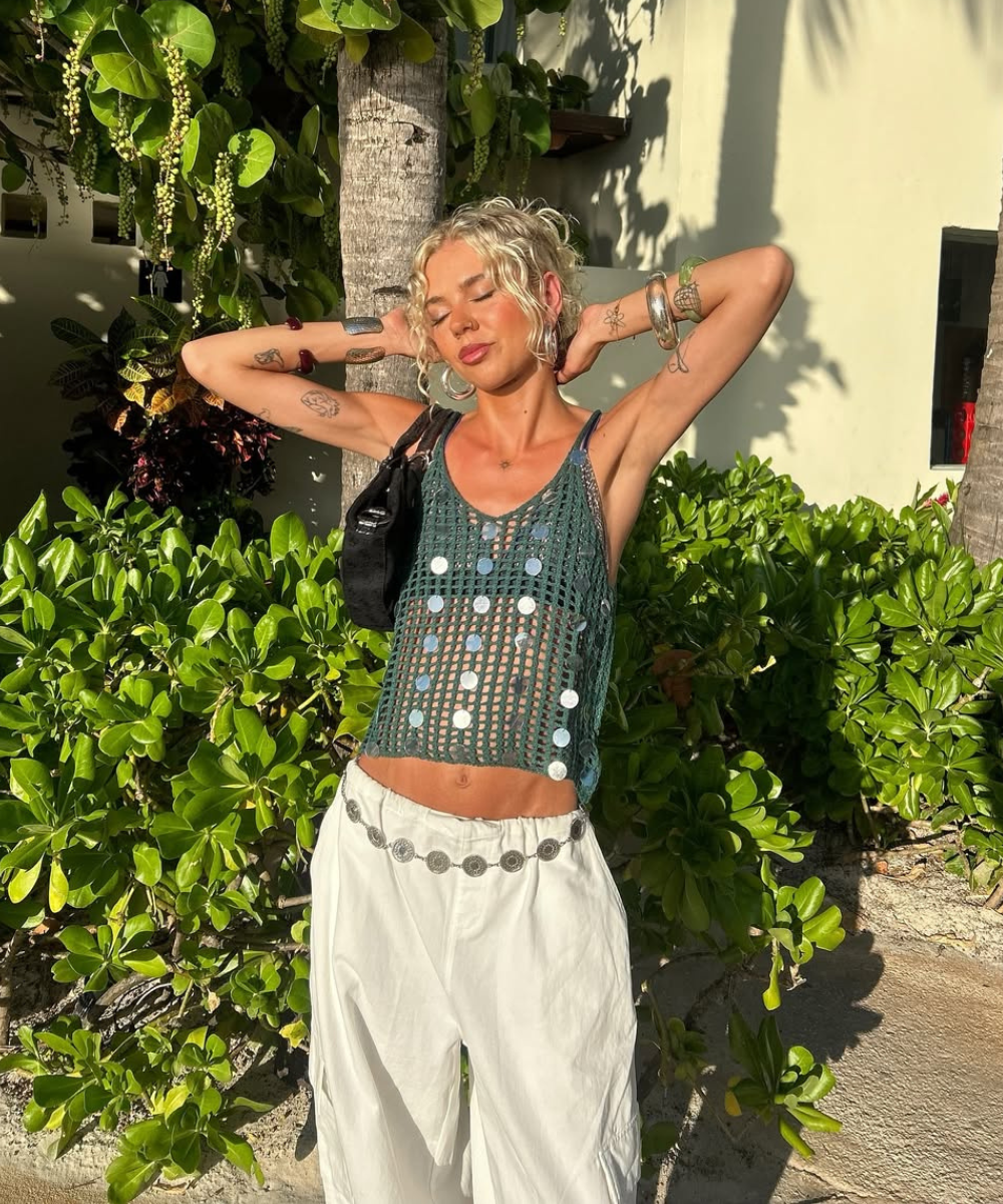 Pessoa em look casual, com blusa rendada e calça branca, posa sob o sol ao lado de plantas.