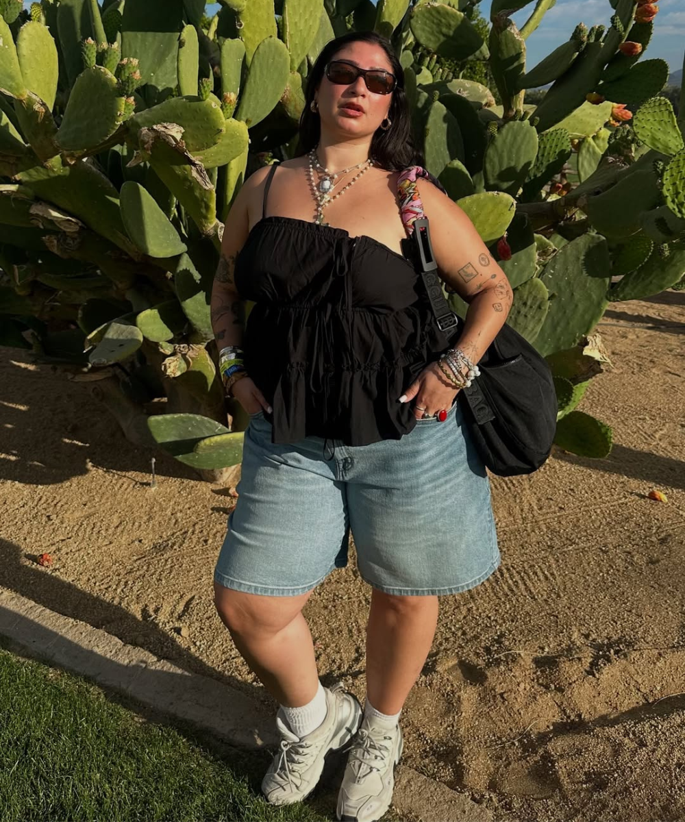 Pessoa em frente a cactos, usando blusa preta, bermuda jeans e tênis. Acessórios são parte do look.