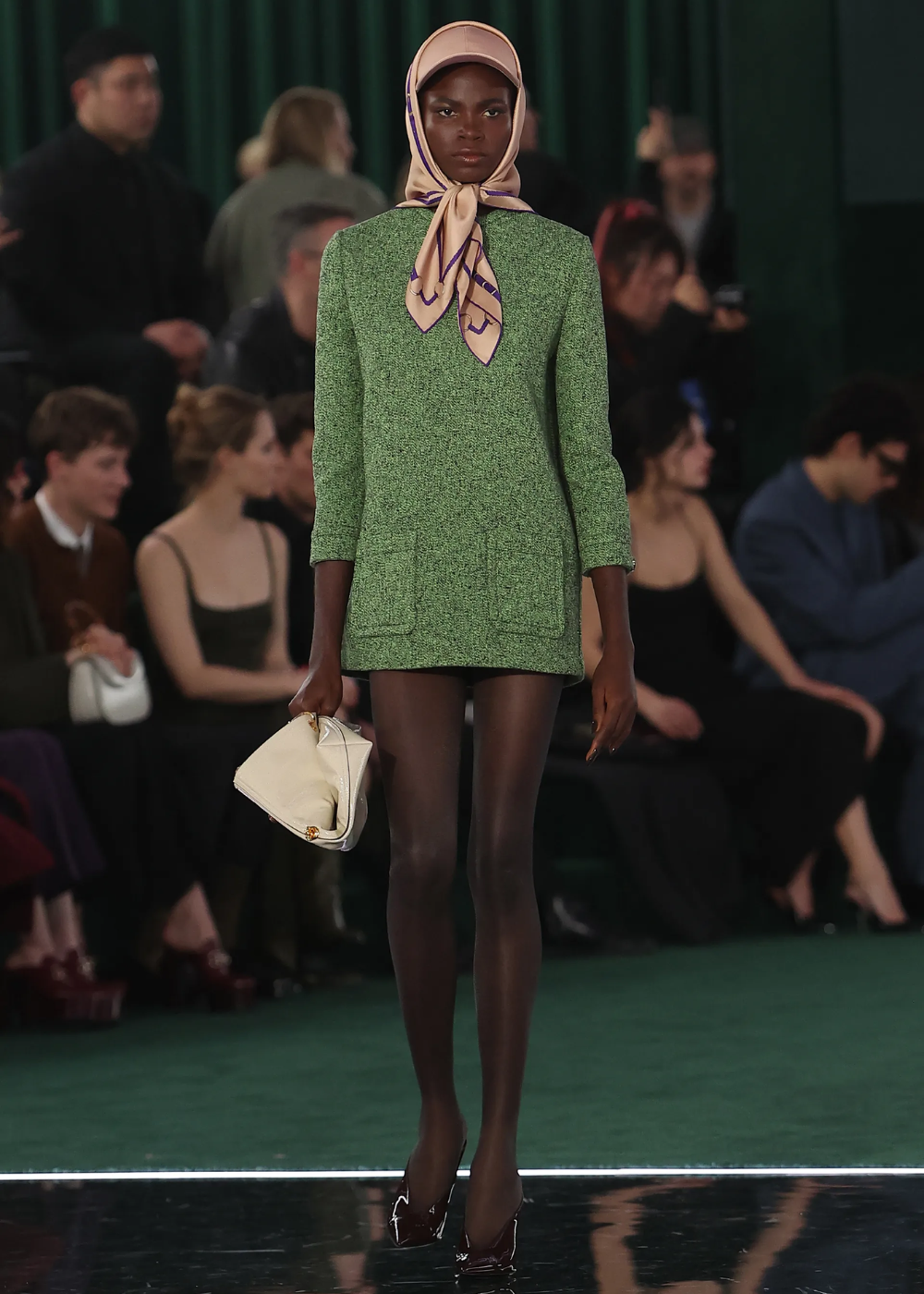 Modelo em passarela usa vestido verde curto e lenço de seda. Acessórios de marcas desejadas. Público ao fundo.