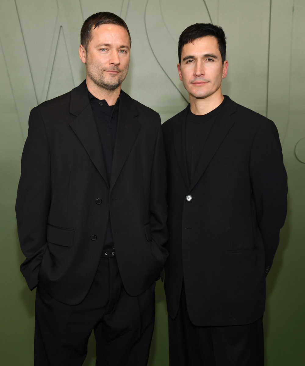 Dois homens posam com ternos escuros contra um fundo verde, transmitindo elegância.