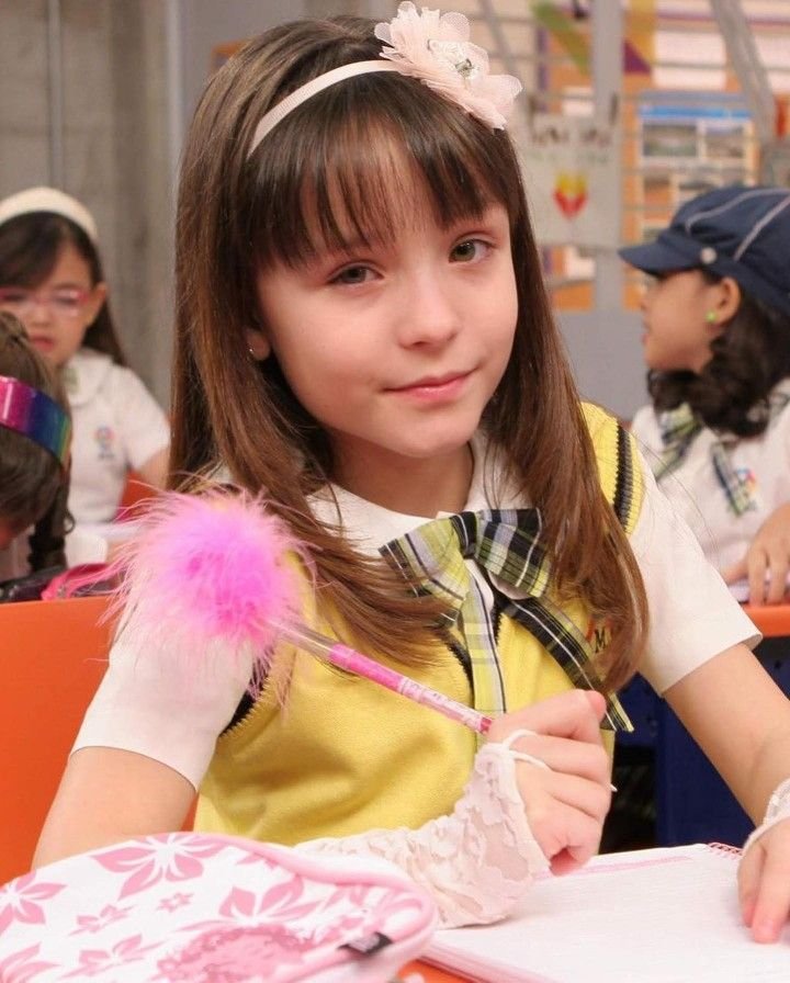 Menina com faixa de cabelo e uniforme escolar segura caneta com pompom rosa em sala de aula. Ideal para cenas de novelas.
