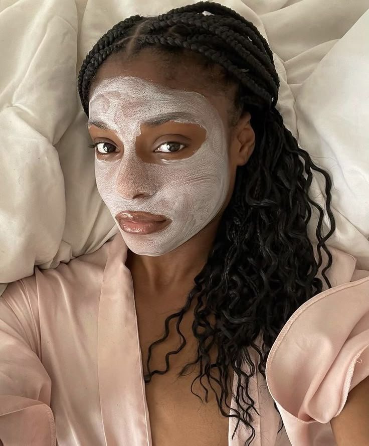 Mulher com uma máscara facial caseira branca relaxa em uma cama, usando um roupão rosa claro.