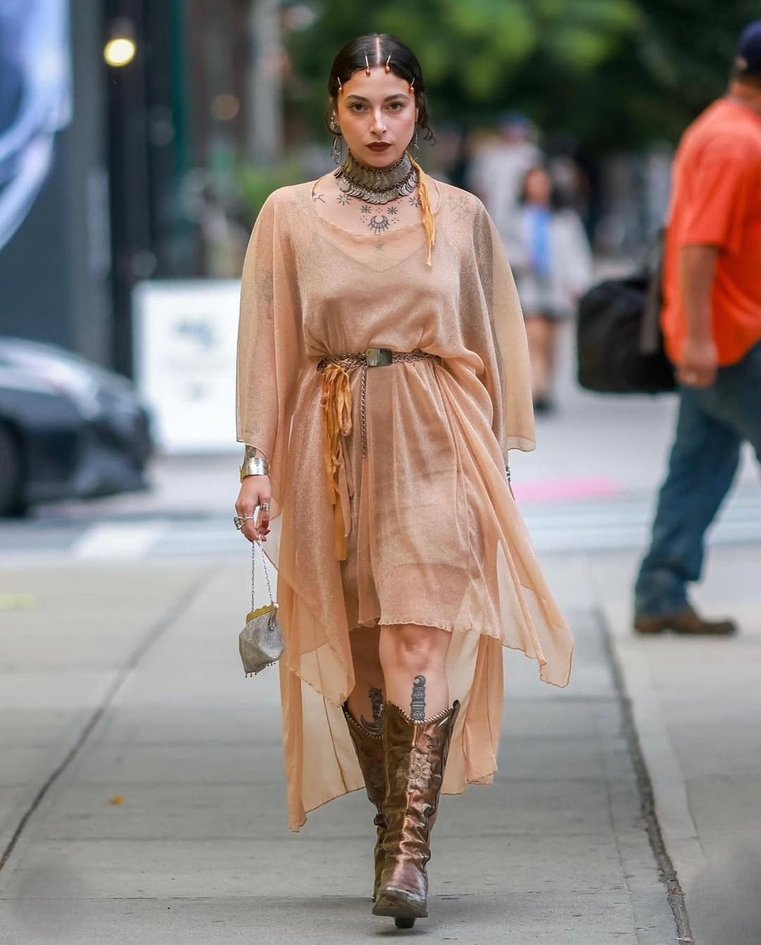 Pessoa veste traje fluido e acessórios pesados em estilo medieval urbano, caminhando em uma rua movimentada.