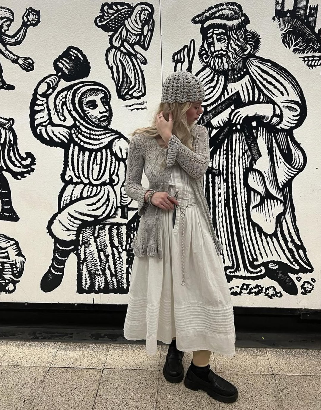 Pessoa com roupas de estilo boêmio posando em frente a mural medieval em preto e branco. Medieval core