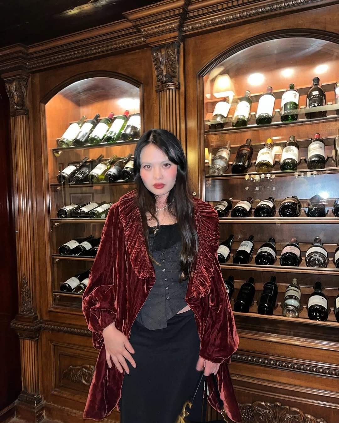 Pessoa vestindo roupa vintage em frente a adega com garrafas de vinho em armário de madeira decorado.